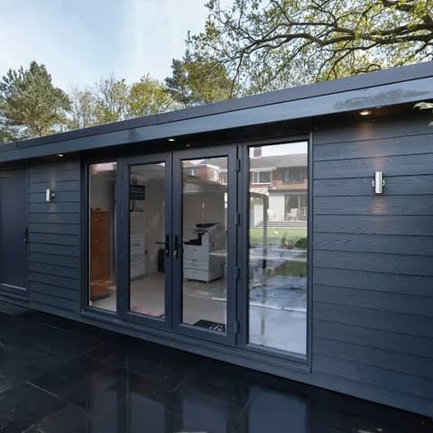 Modern dark blue garden room or studio with glass doors and windows, set in a residential yard with trees in background