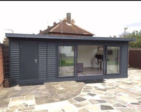 Modern dark gray garden studio with large glass doors and patio, set against a brick house with tiled roof