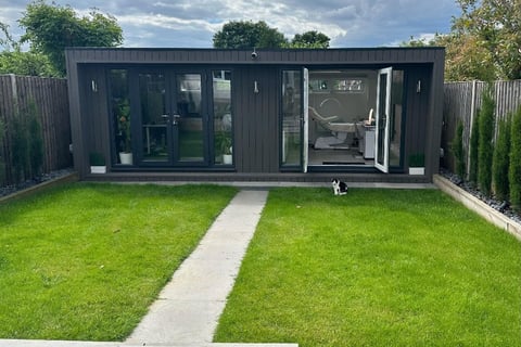 Modern black garden studio with large windows, concrete pathway, and manicured lawn surrounded by wooden fencing