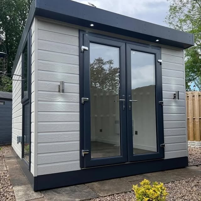 Modern garden studio with grey horizontal siding, black frame, and large glass double doors on a gravel patio