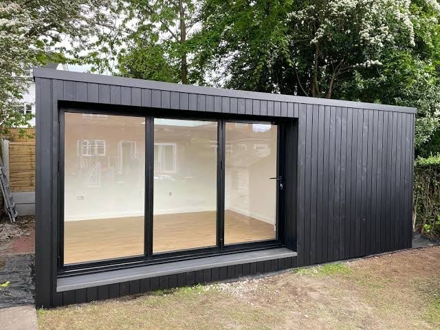 Modern black garden studio with large sliding glass doors, wooden floor, and corrugated metal exterior in landscaped yard