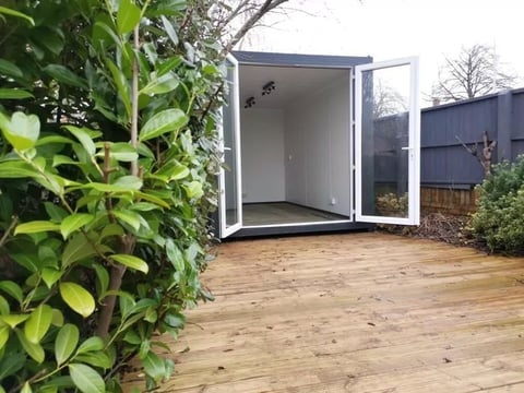 Modern white and dark gray garden shed with open double doors on wooden deck, surrounded by green foliage and landscaping